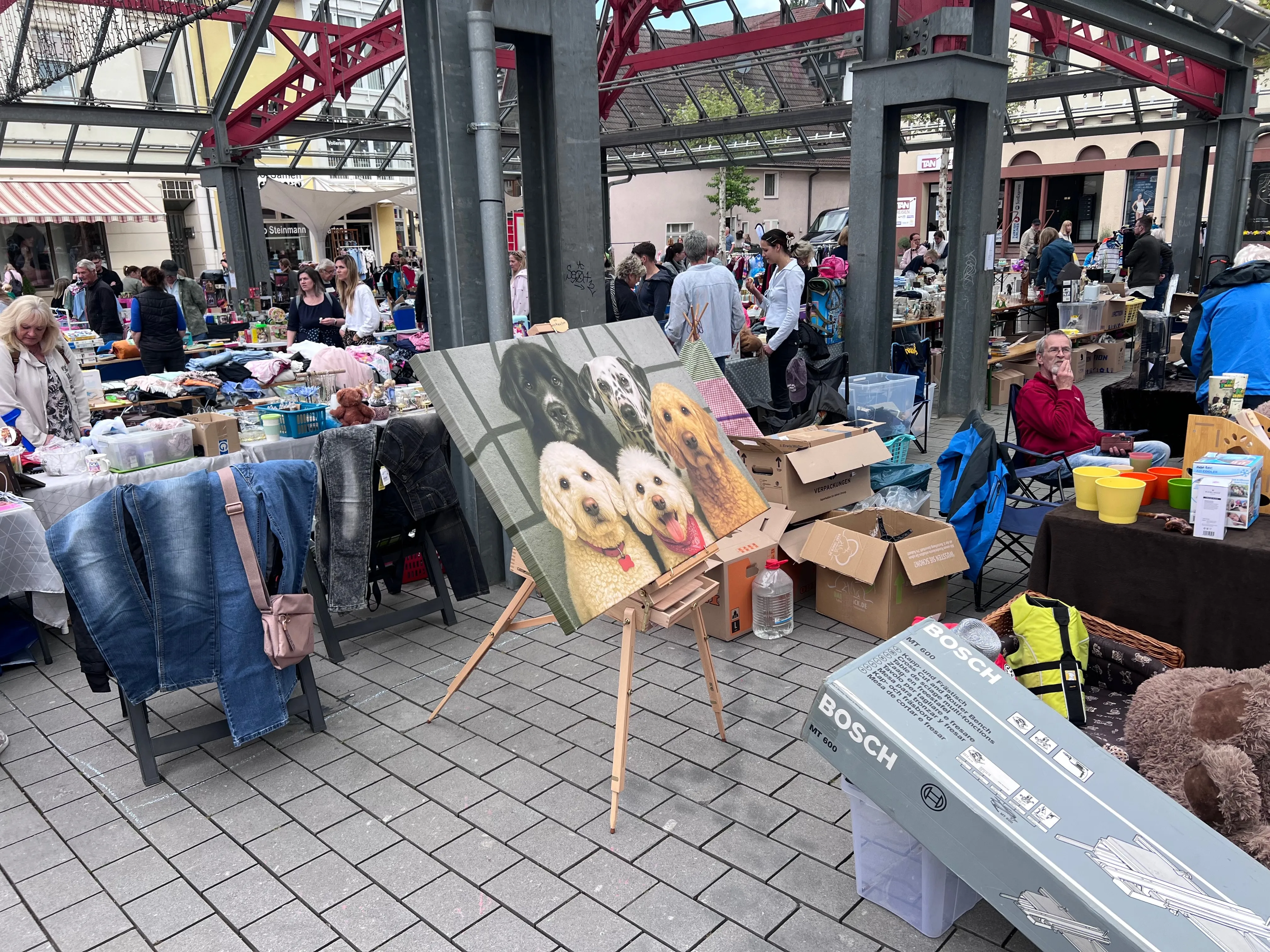 Ein belebter Frühlingsmarkt in einer Innenstadt mit vielen Menschen, Ständen und einem Künstler, der ein Porträt von zwei Hunden malt.