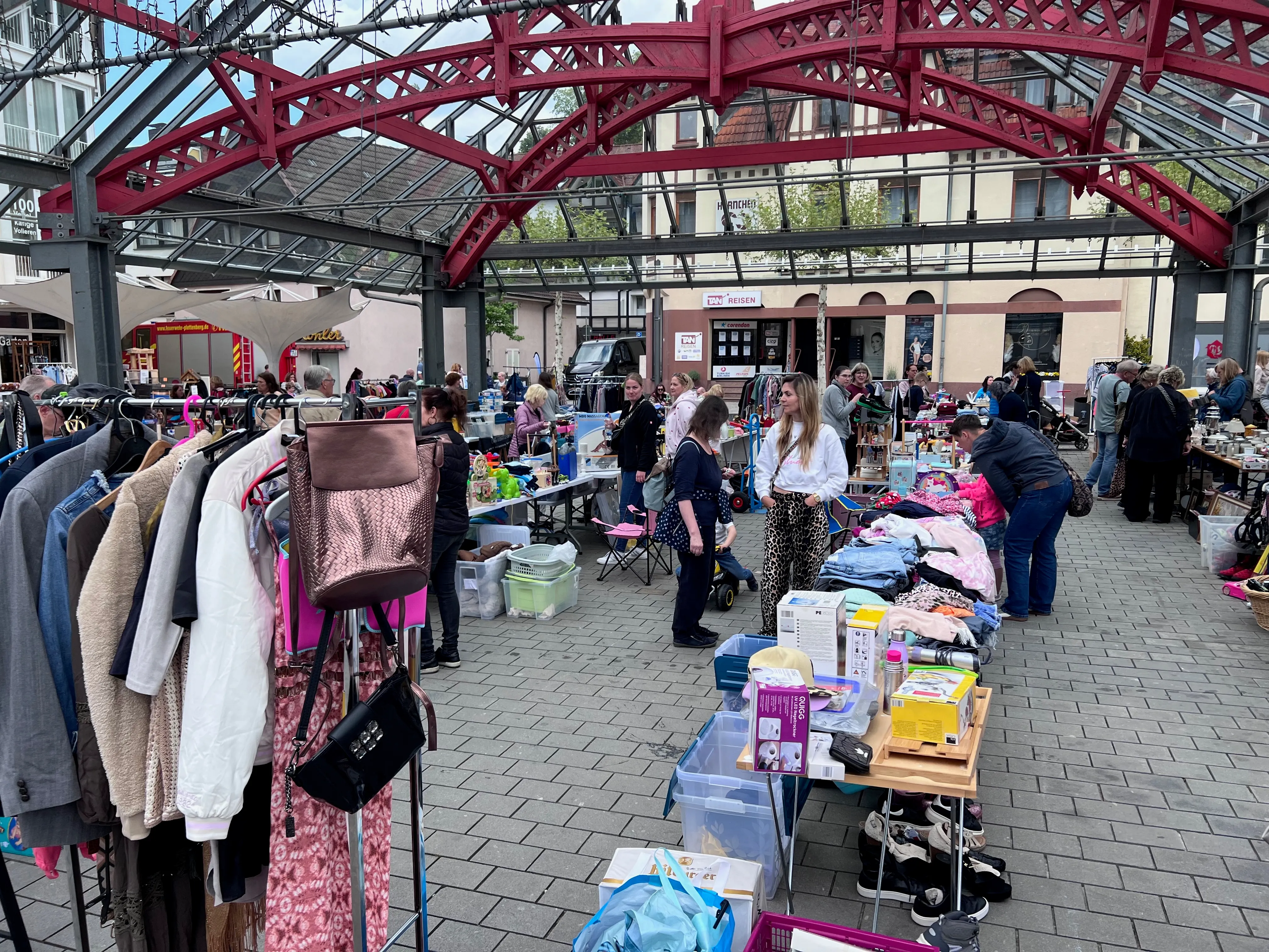 Ein belebter Frühlingsmarkt in einer Innenstadt mit Ständen voller Kleidung und Waren, umgeben von Besuchern aller Altersgruppen.