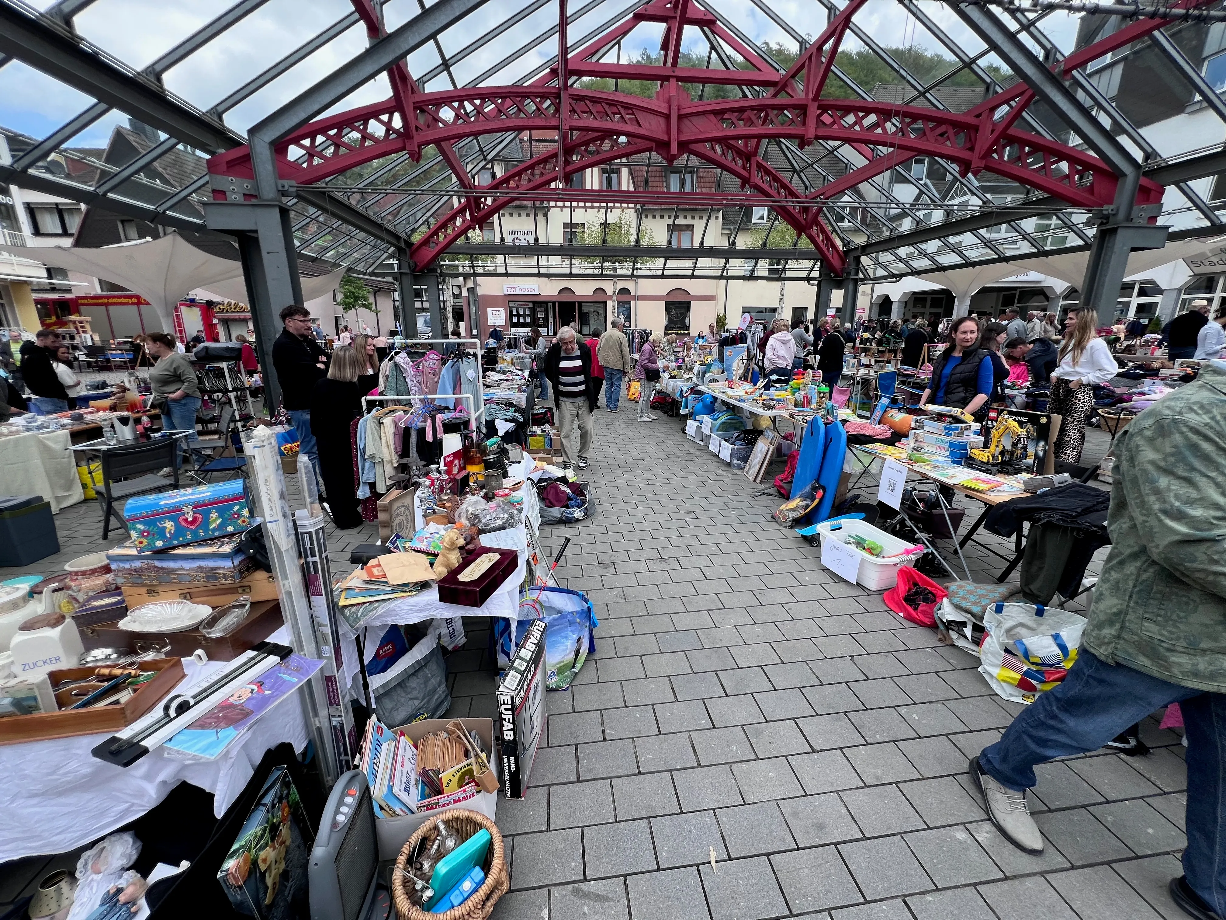 Blick durch eine Glasüberdachung auf einen belebten Flohmarkt in einer Innenstadt. Menschen stehen an Tischen mit verschiedenen Artikeln, im Hintergrund ein Gebäude mit weiteren Marktständen.