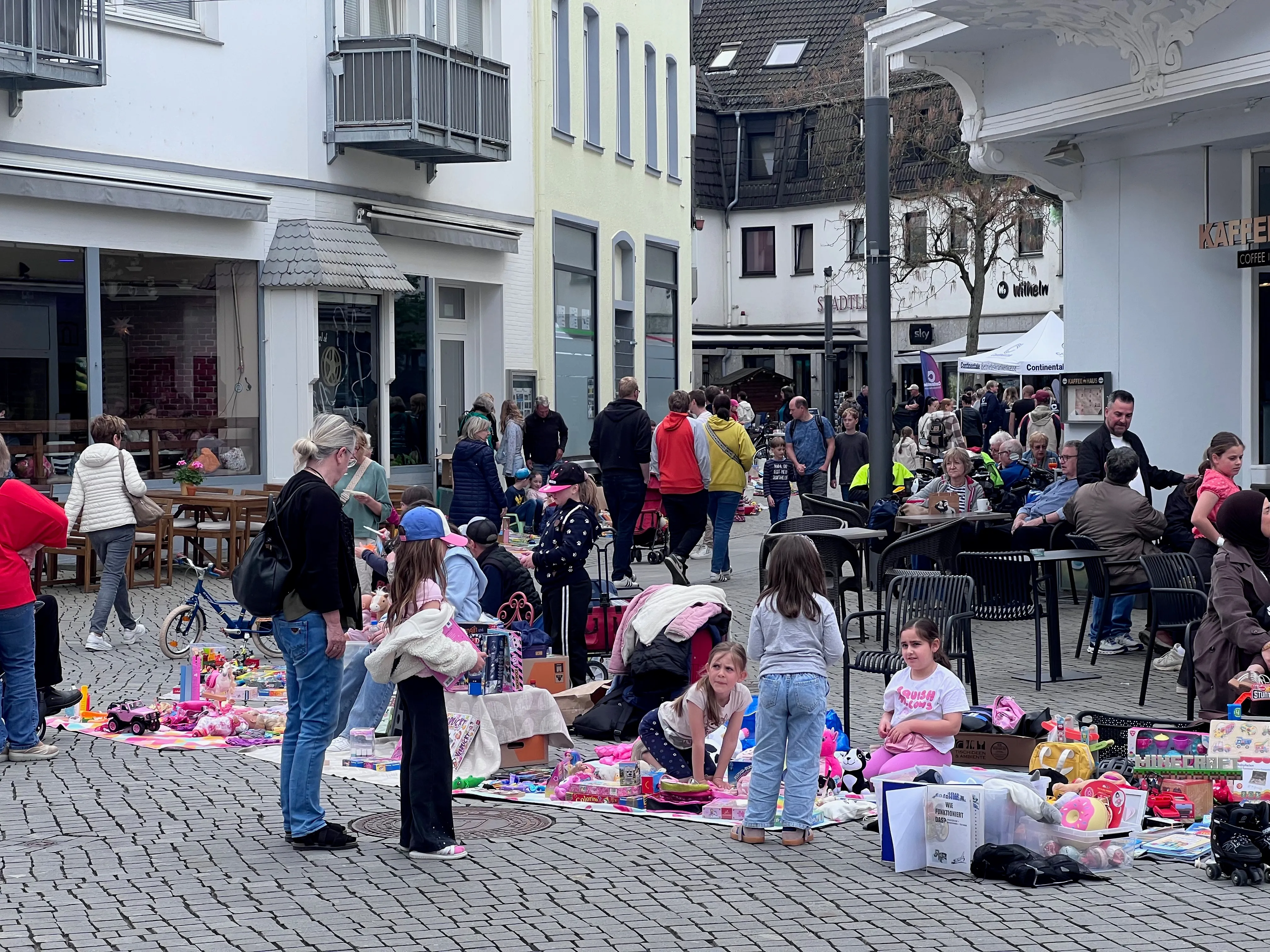 Ein belebter Platz in einer Innenstadt ist gefüllt mit Menschen, die einen Flohmarkt besuchen. Kinder stehen vor Tischen mit Spielzeug, während Erwachsene stöbern und sich unterhalten.