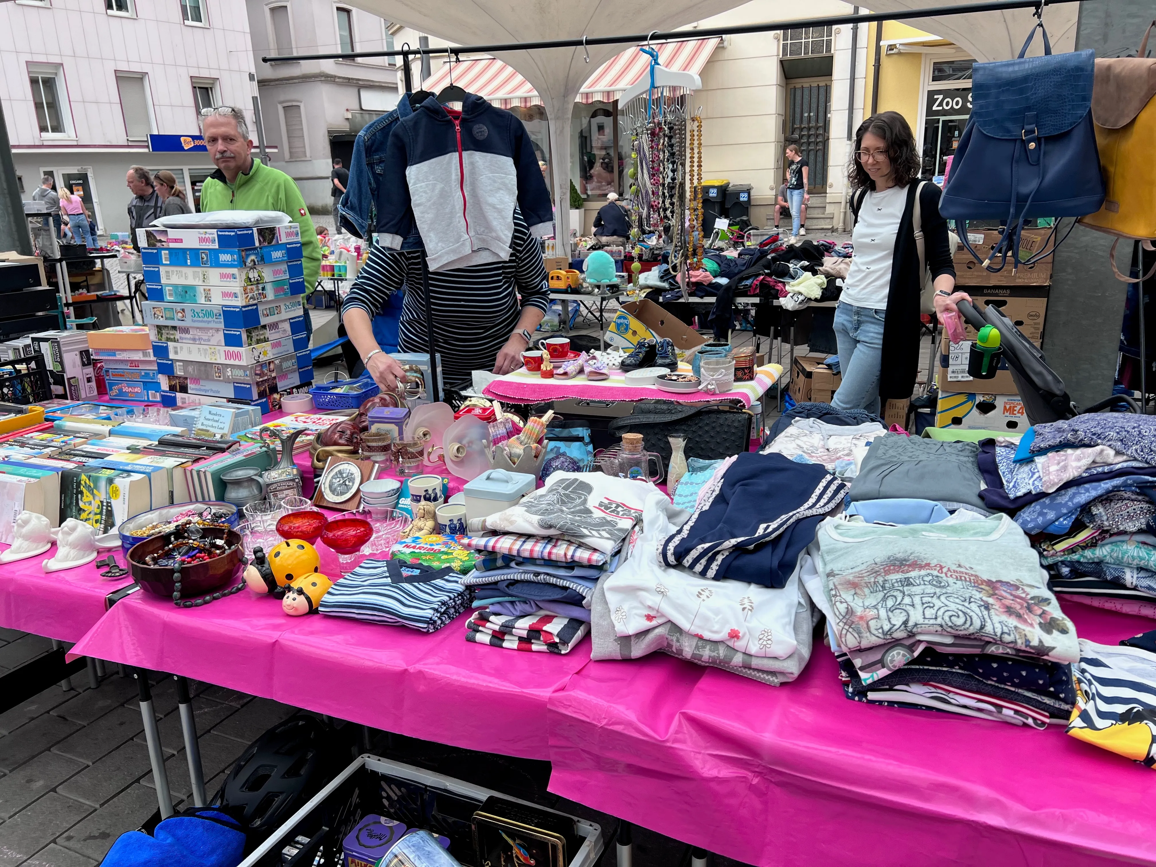 Ein belebter Stand auf einem Flohmarkt mit Kleidung, Spielzeug und anderen Gegenständen. Im Hintergrund stehen Menschen und stöbern.