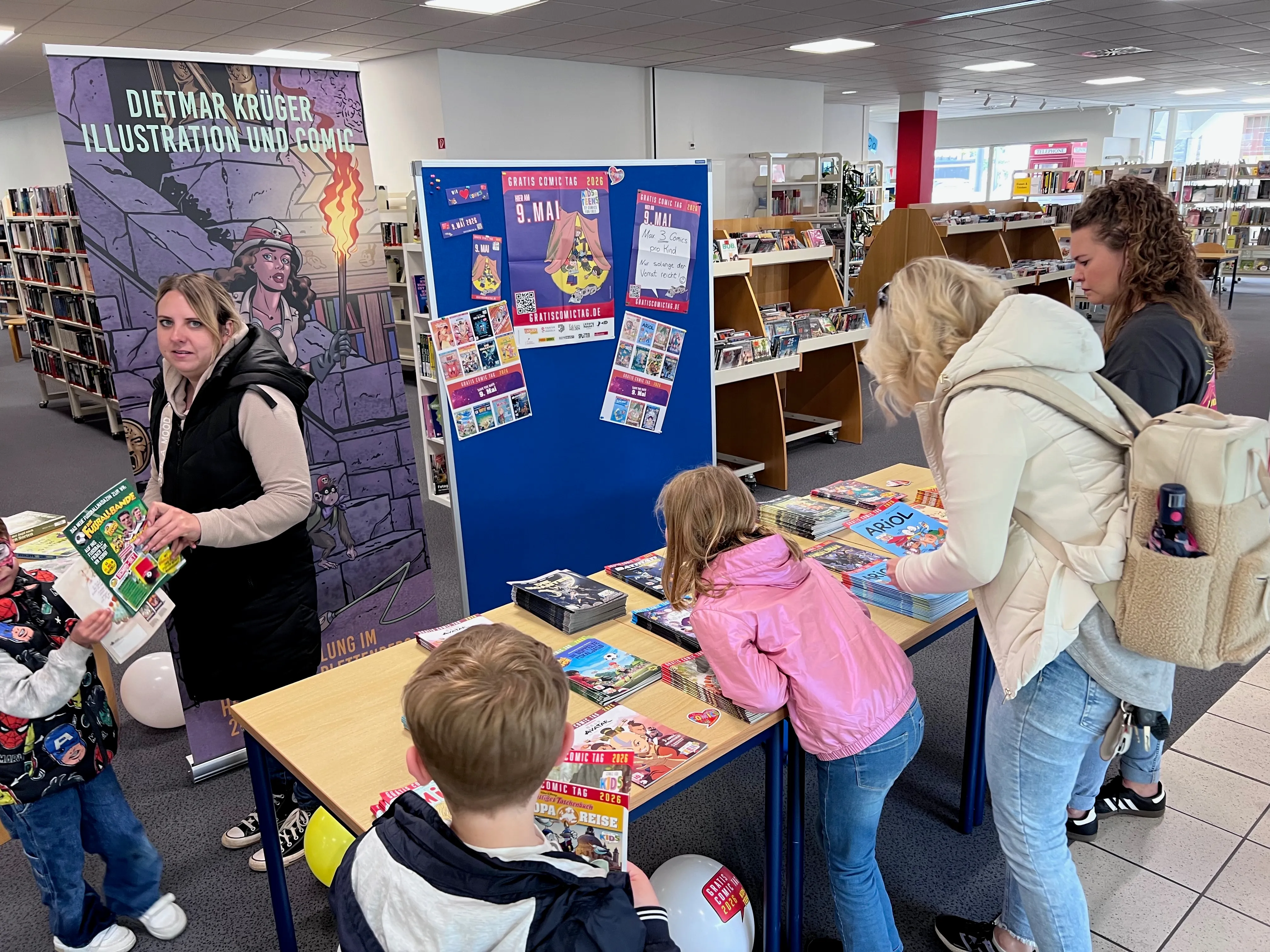 Comiczeichnerin sitzt an einem Tisch mit Kinderbüchern und unterzeichnet ein Exemplar für ein Kind, während andere Kinder und Eltern in der Bücherei stehen und Comicbücher auswählen.