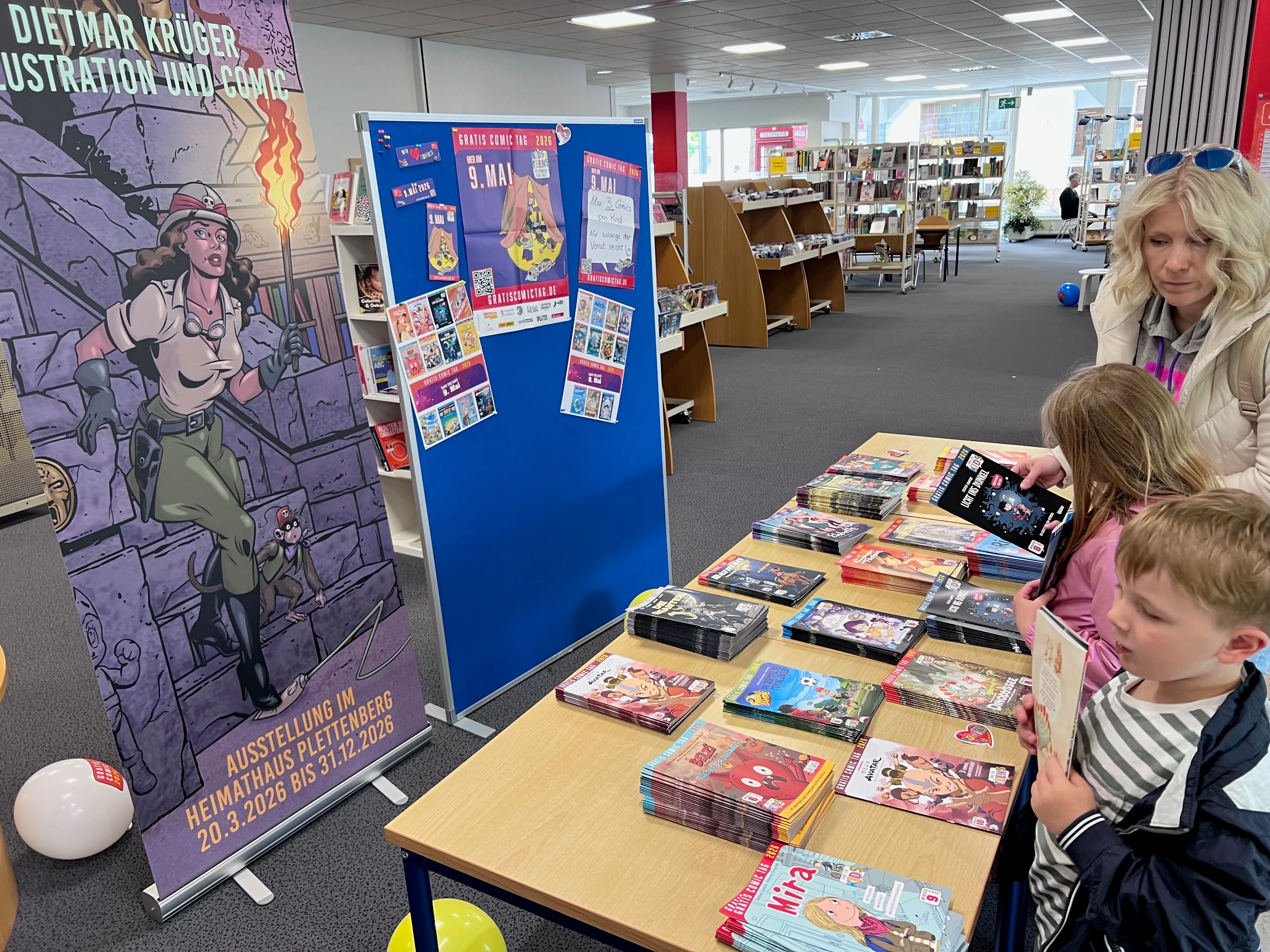 Zwei Kinder betrachten Comicbücher auf einem Tisch in der Stadtbücherei, während im Hintergrund eine Frau steht. Ein Roll-up-Banner mit Comiczeichnungen und Informationen zum kostenlosen Comic-Tag ist zu sehen.