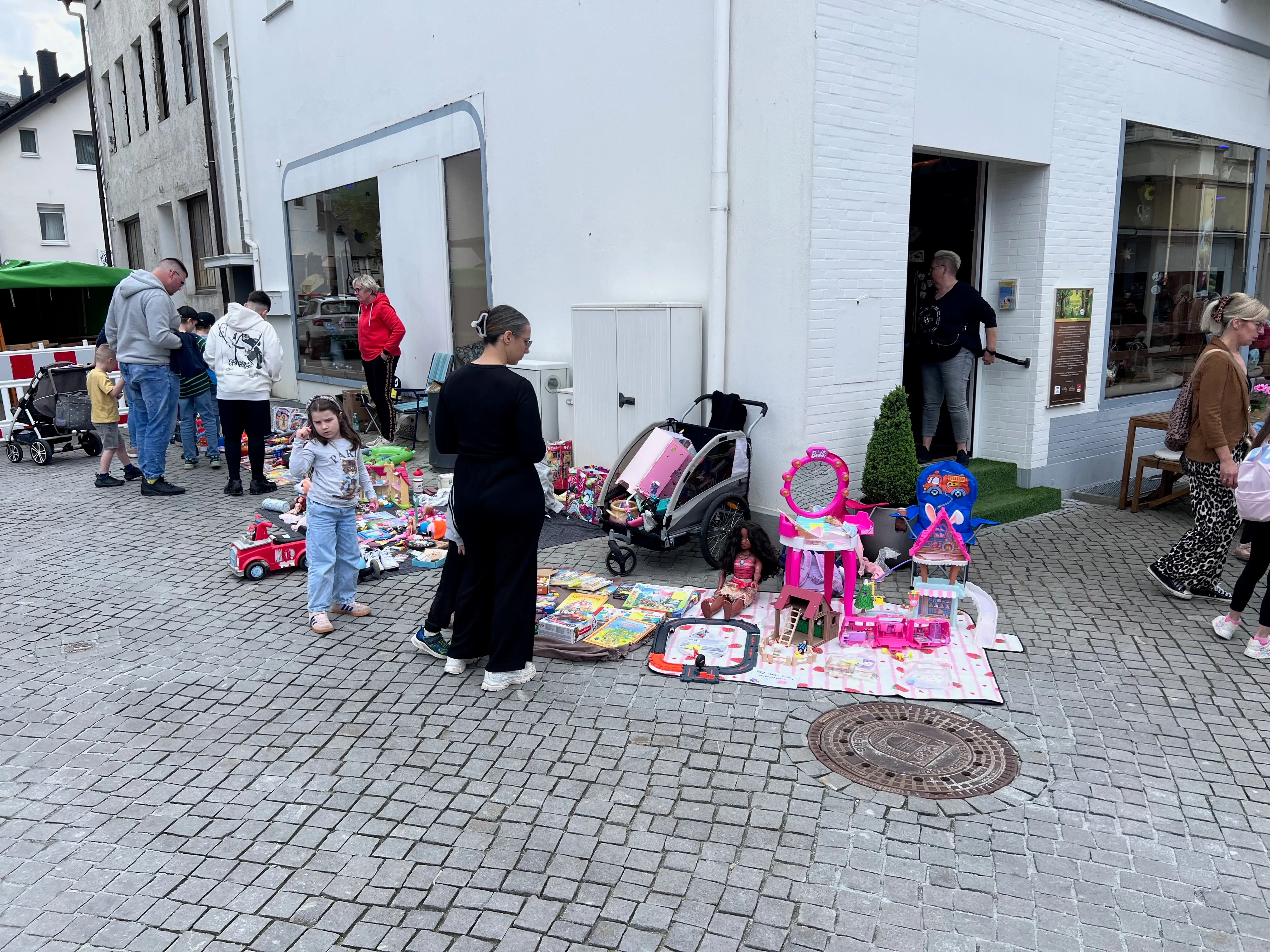 Menschen auf einem Flohmarkt in einer Fußgängerzone, mit Spielzeug und Kleidung auf Tischen und Decken ausgebreitet.