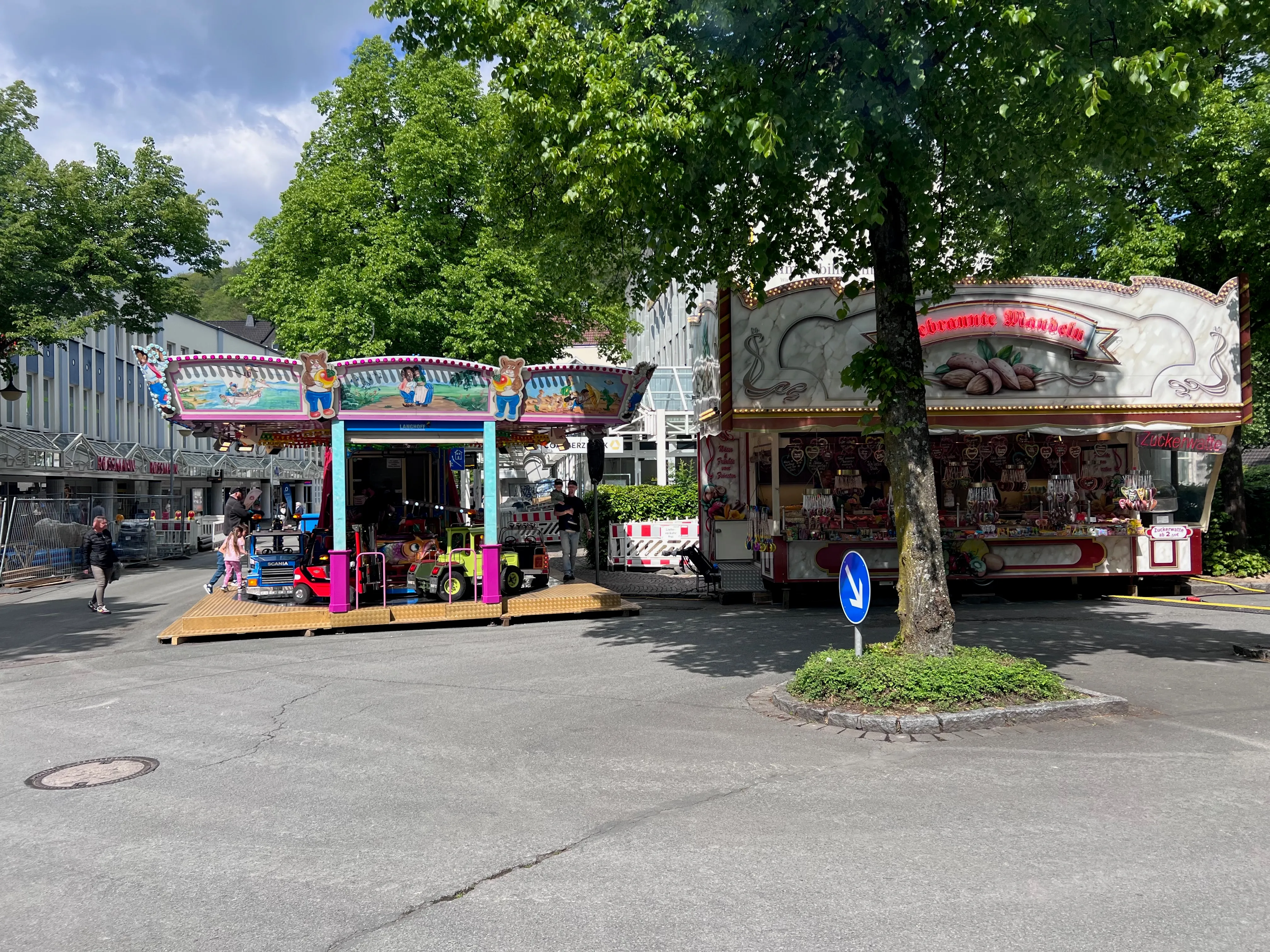 Ein bunt geschmückter Karussellstand und ein Süßigkeitenstand stehen auf einem Pflasterplatz vor Bäumen. Im Hintergrund sind weitere Marktstände und Menschen zu sehen.
