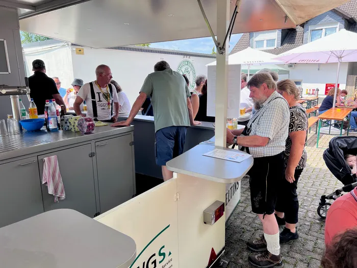 An einer mobilen Bar bedienen zwei Personen Gäste mit Bier. Im Hintergrund stehen und sitzen weitere Menschen, einige mit Getränken, vor einem Schützenverein.