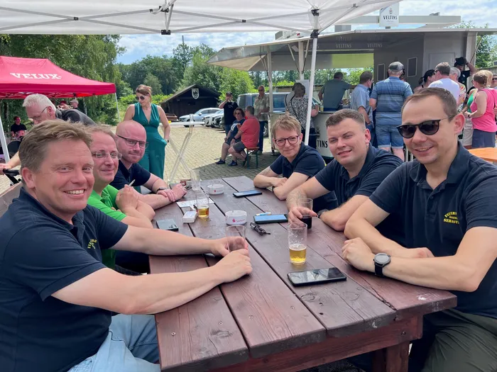 Fünf Männer sitzen an einem Holztisch im Freien, Biergläser und Smartphones stehen davor. Im Hintergrund sind weitere Menschen und Zelte zu sehen, die zu einem Schützenfest gehören.