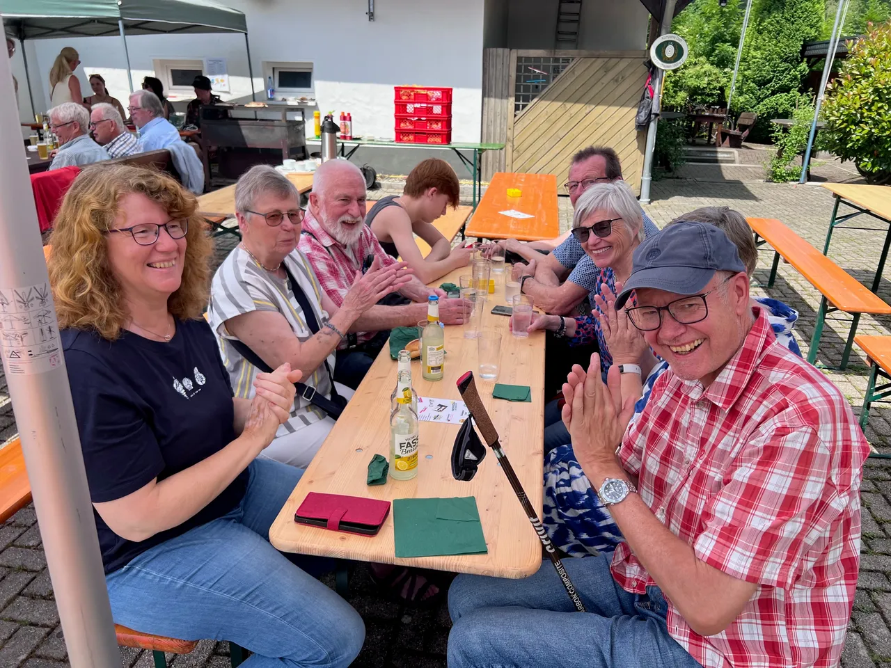 Eine Gruppe von sieben Menschen sitzt an einem Holztisch im Freien, lächelt und scheint sich zu unterhalten. Auf dem Tisch stehen Getränke und ein Tablett mit Snacks. Im Hintergrund sind weitere Personen und ein Gebäude zu sehen.