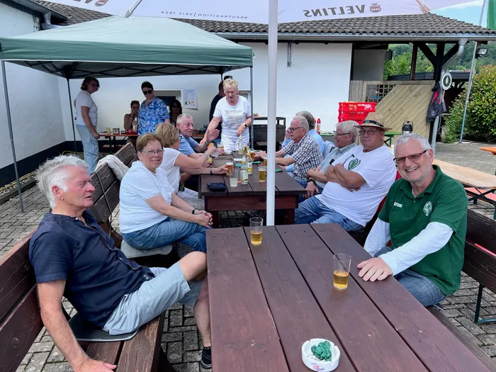 Eine Gruppe von Menschen sitzt an Biertischen vor einem Gebäude und trinkt Bier. Einige tragen Kleidung mit einem Vereinslogo.