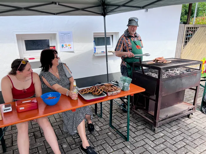 Ein Mann brät Würstchen auf einem Holzkohlegrill unter einem Pavillon. Zwei Frauen sitzen an einem Tisch davor, eine davon hält ein Glas Bier. Im Hintergrund ist ein Plakat zu sehen.