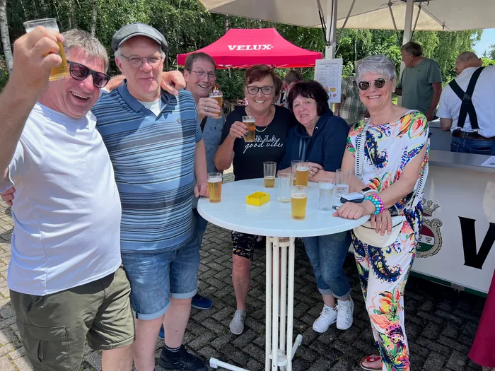Eine Gruppe von sechs Personen steht um einen runden Tisch und hält Biergläser hoch. Im Hintergrund ist ein roter Pavillon und ein Getränkestand zu sehen.