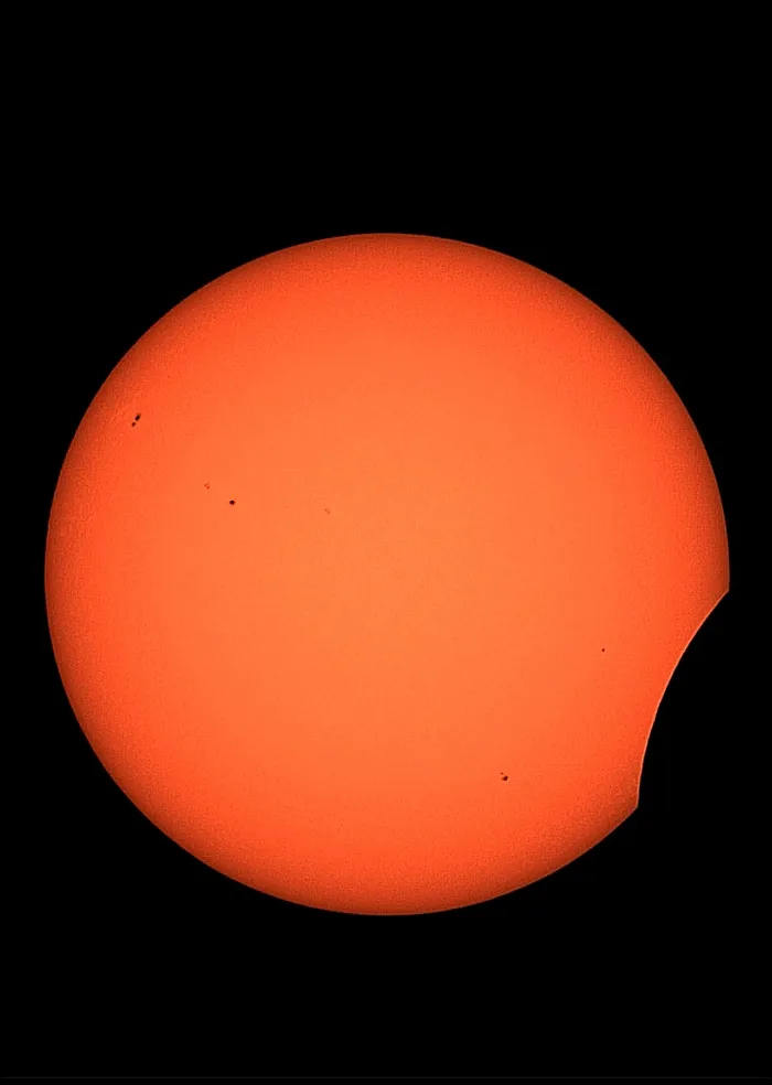 Leuchtend orangefarbene Sonne mit sichtbaren Sonnenflecken, die teilweise von der dunklen Silhouette des Mondes verdeckt wird.