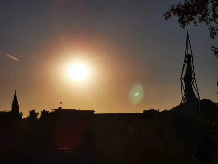 Die Sonne und eine Mondsichel am Abendhimmel über der Silhouette einer Stadt mit markanten Türmen.