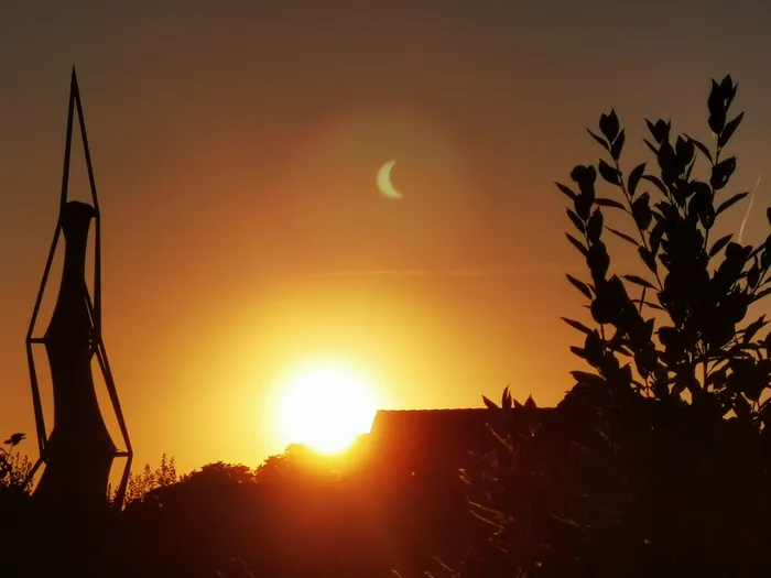Silhouette einer Skulptur und von Bäumen vor einem orangefarbenen Himmel mit der Sonne und einer Mondsichel.