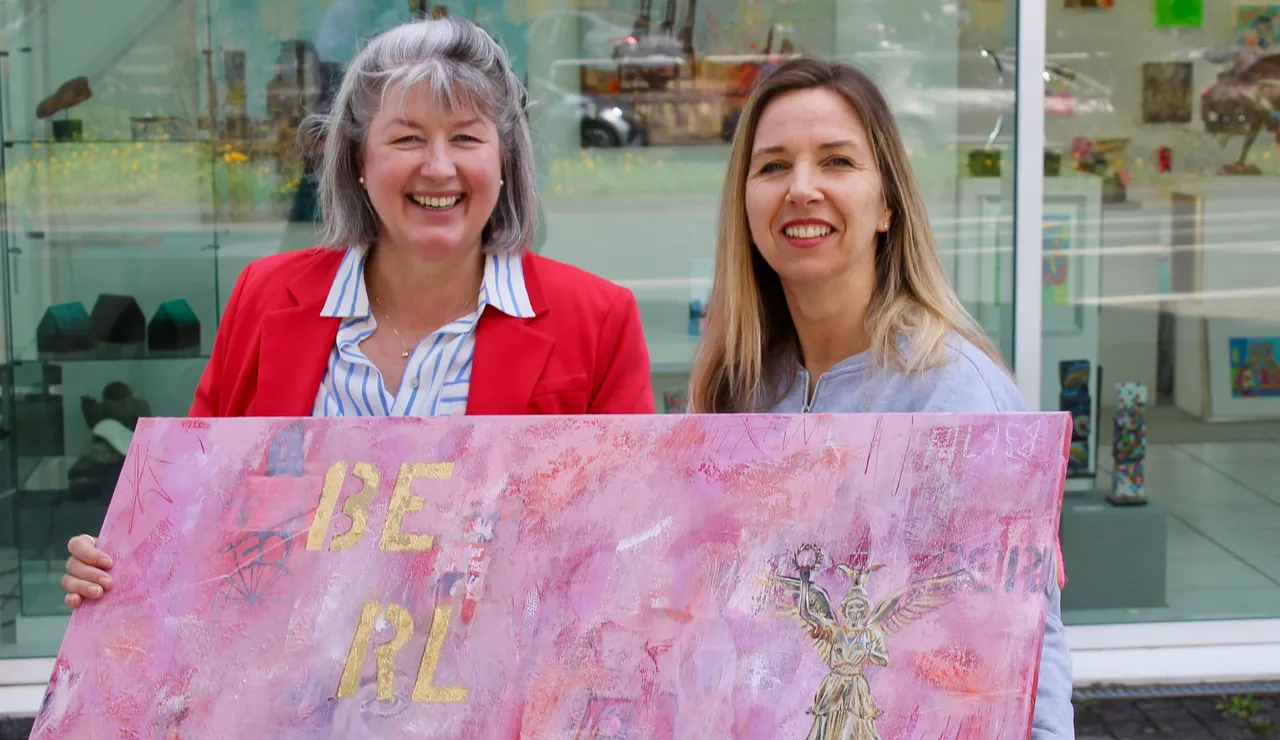 Zwei lächelnde Frauen, Claudia Ackermann und Galeristin Nadja Schmidt-Widua, halten ein Gemälde mit dem Schriftzug „BE REAL“ vor einer Galerie.