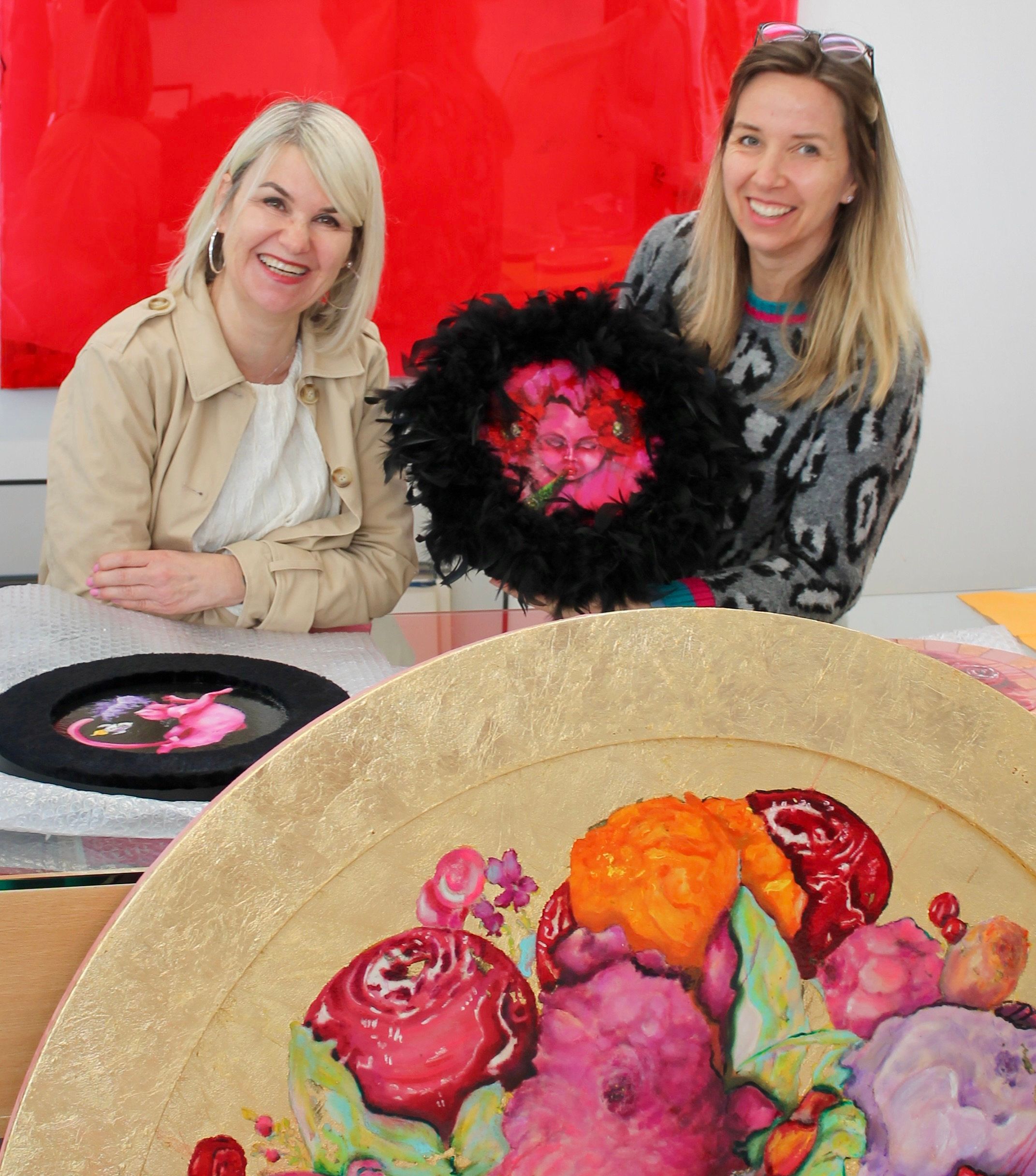 Zwei lächelnde Frauen sitzen vor ihren Gemälden. Im Vordergrund ein farbenfrohes, rundes Bild mit floralen Motiven.