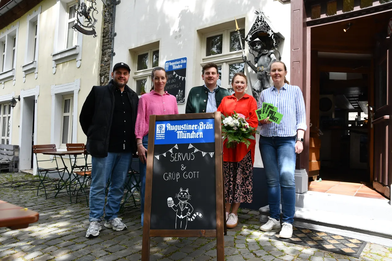 Fünf Personen stehen vor einem Restaurant mit dem Namen „Kater August“. Ein Schild mit einer Katze und der Aufschrift „Servus & Grüb Gott“ steht davor.