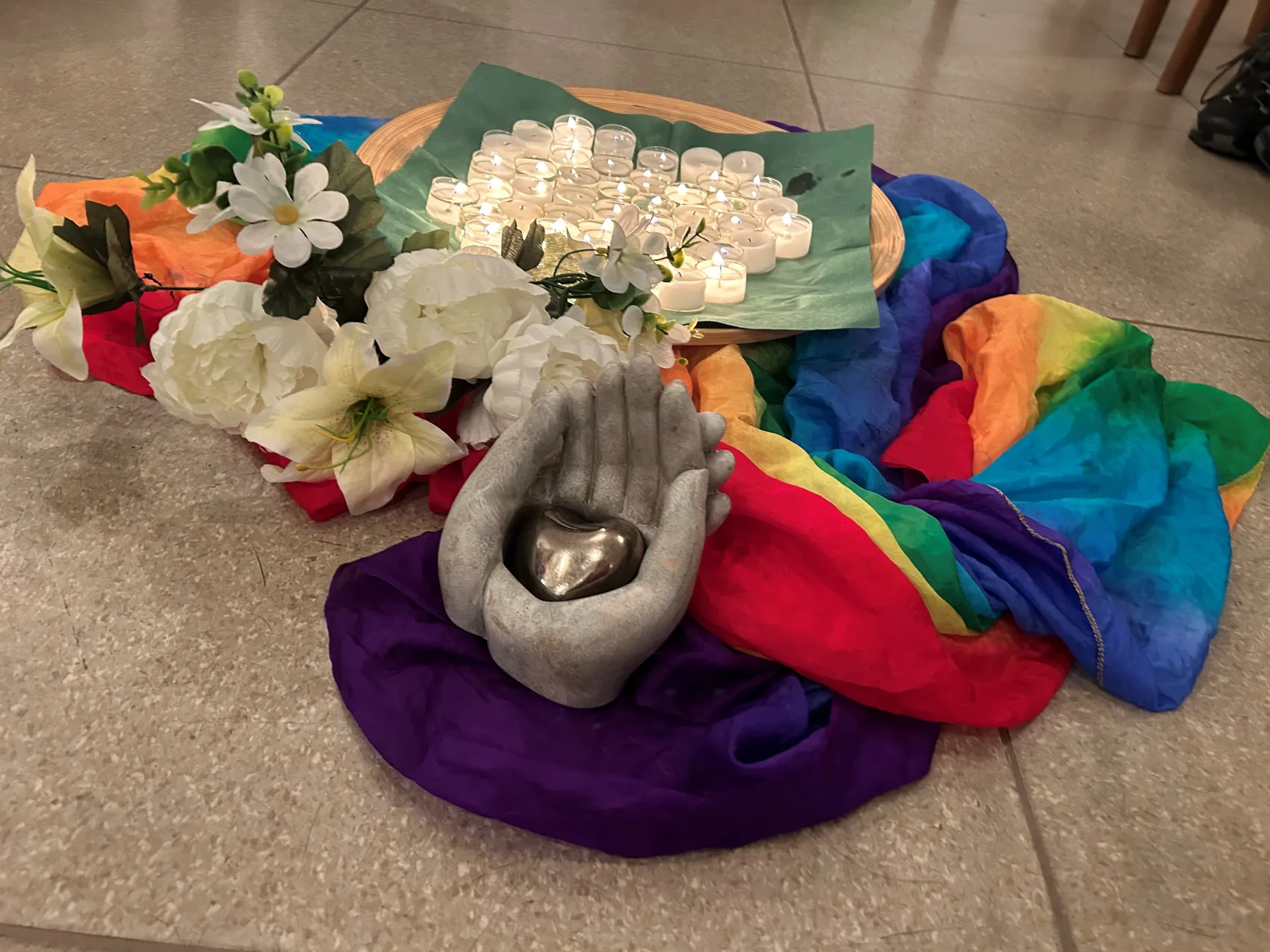 Eine steinerne Hand hält ein goldenes Herz, umgeben von weißen Blumen, Kerzen und einem Regenbogen-Tuch. Das Bild symbolisiert Trauer, Verlust und Unterstützung.
