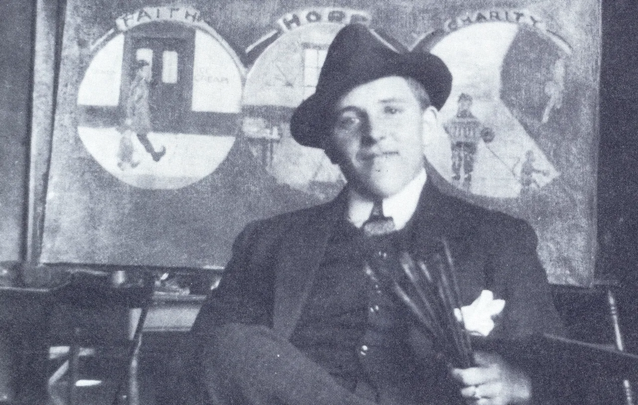 Ernst Geitlinger in Anzug und Hut vor einem Gemälde mit den Motiven „Faith“, „Hope“ und „Charity“.
