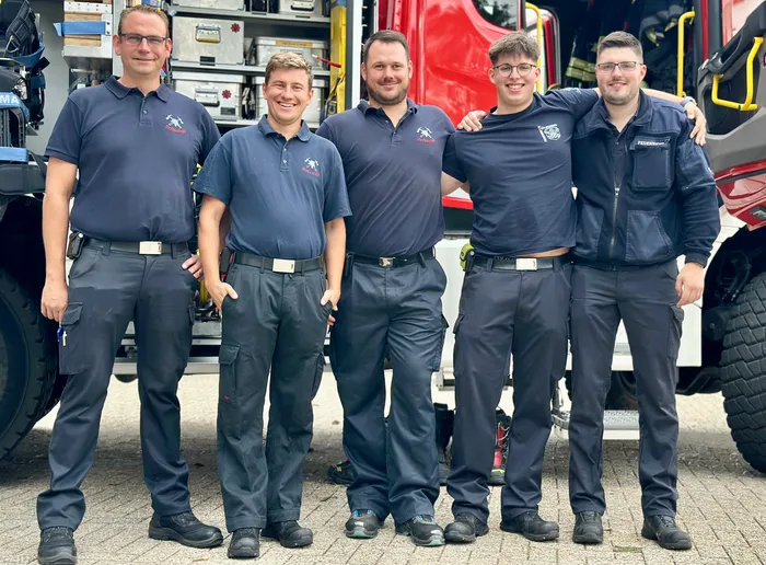 Fünf Männer in dunkelblauen Uniformen stehen lächelnd vor einem roten Feuerwehrfahrzeug.
