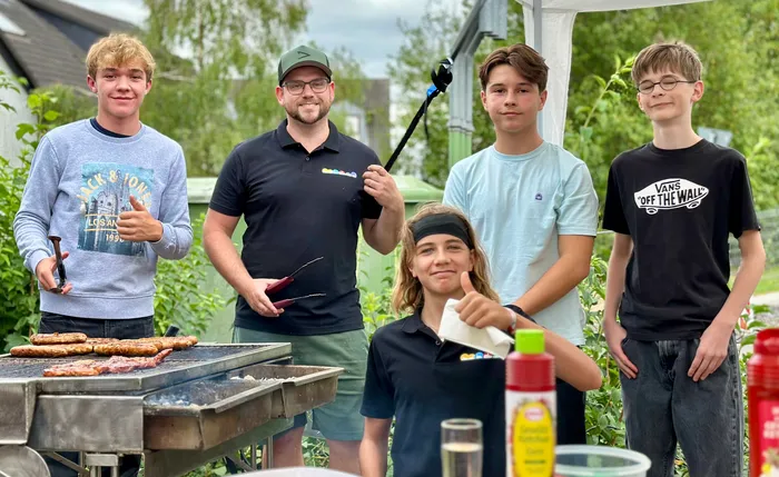 Fünf junge Männer stehen hinter einem Grill mit Bratwürsten und Fleisch bei einem Gemeindefest.