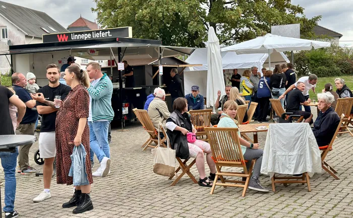 Besucher sitzen an Holztischen unter weißen Sonnenschirmen und stehen vor einem Imbisswagen.