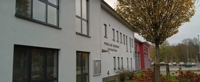 Das weiße Gebäude der Freiwilligen Feuerwehr Löschzug Brügge mit mehreren Fenstern und einem Baum davor.