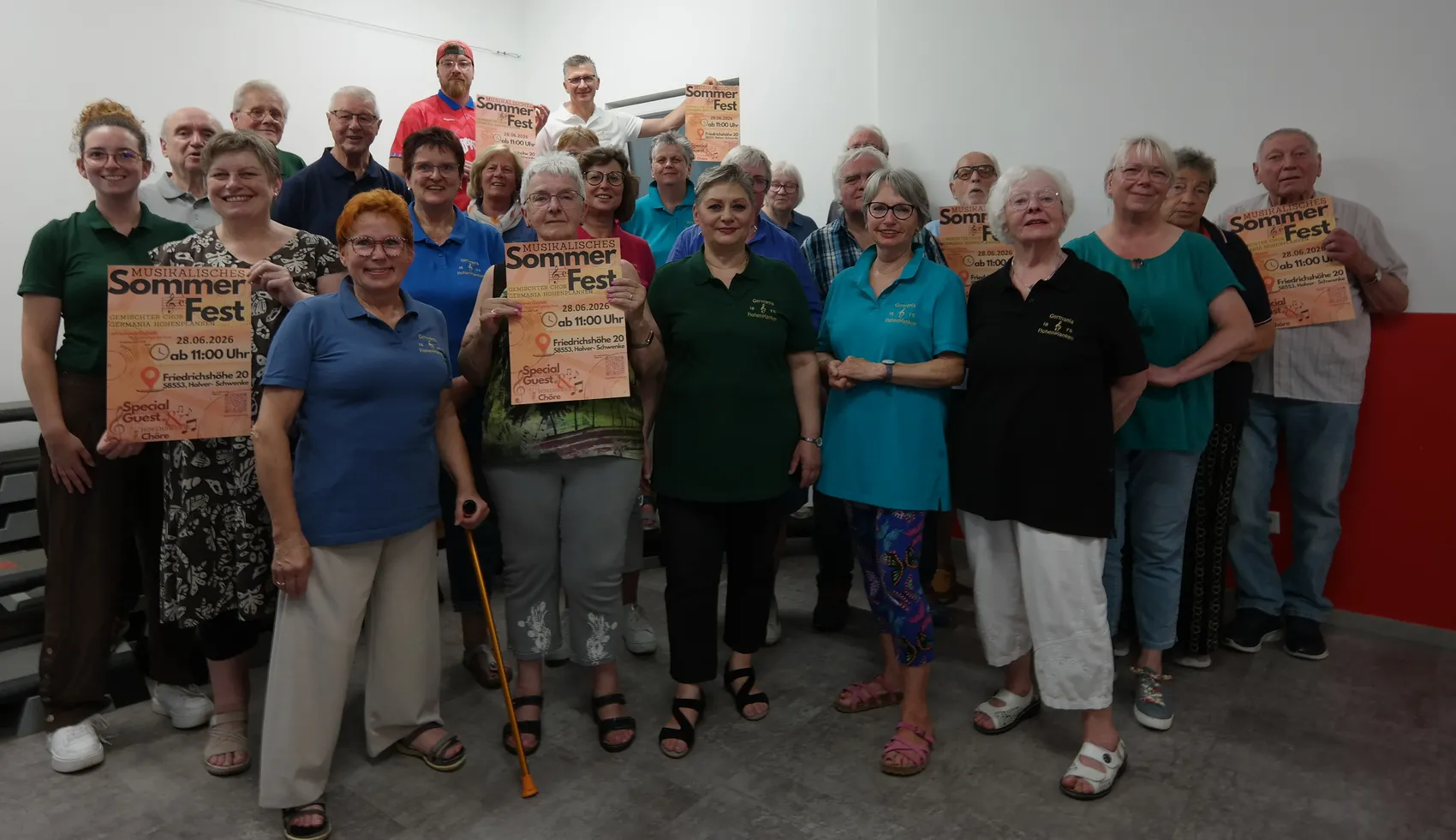 Eine Gruppe von etwa 30 Menschen, hauptsächlich ältere Erwachsene, posiert für ein Gruppenfoto. Viele halten Plakate mit der Aufschrift „Sommerfest“ und dem Datum „28.06.2026“ in den Händen.