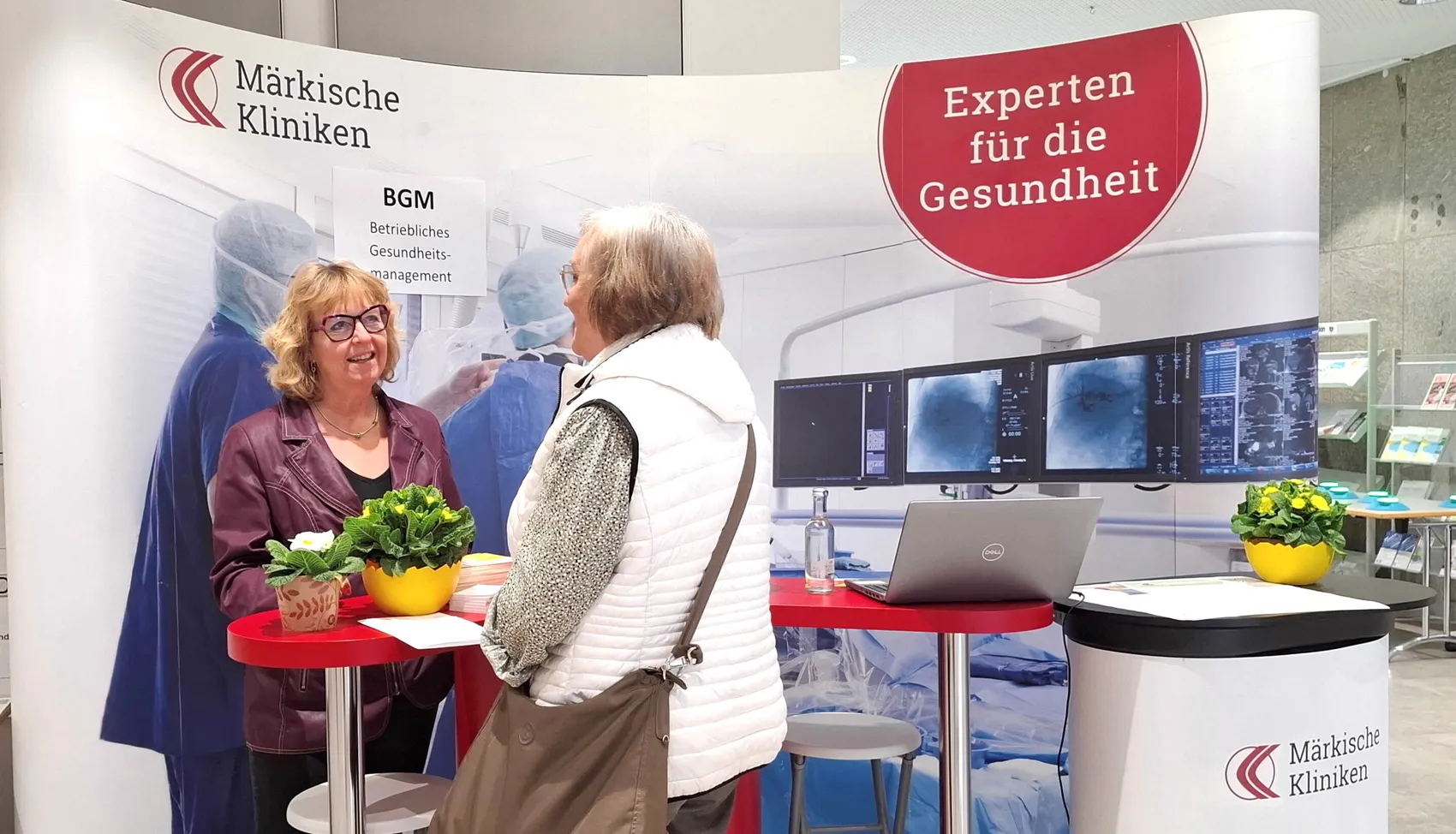 Zwei Frauen stehen an einem Messestand der Märkischen Kliniken vor einem Roll-up-Banner mit dem Aufdruck „Experten für die Gesundheit“ und einem Bildschirm, der medizinische Aufnahmen zeigt.