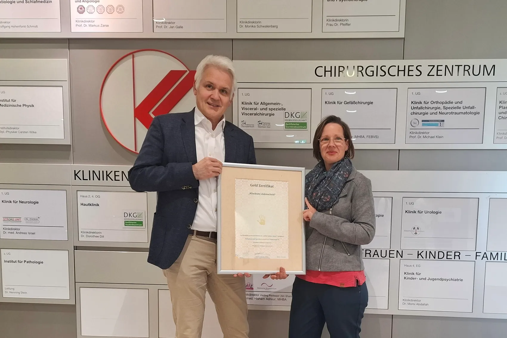 Zwei lächelnde Personen vor einer Wand mit Klinikinformationen und Zertifikaten, darunter ein goldenes Zertifikat mit dem Aufdruck „Exzellentes Maßnahmenpaket“.