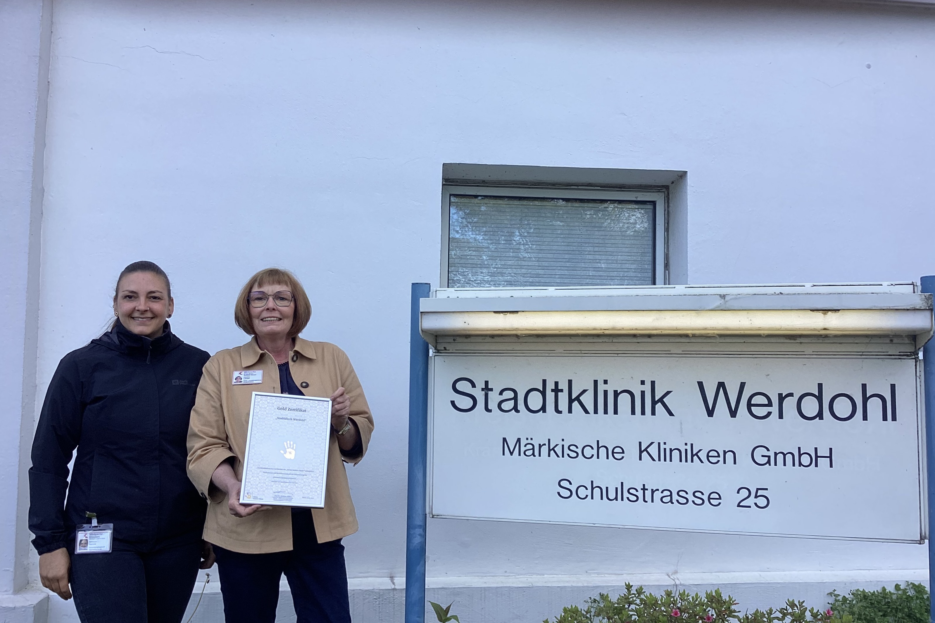 Zwei Frauen vor dem Eingang der Stadtklinik Werdohl halten ein Zertifikat mit einem Handabdruck-Motiv. Die Klinikadresse und der Name der Betreibergesellschaft sind auf einem Schild zu sehen.