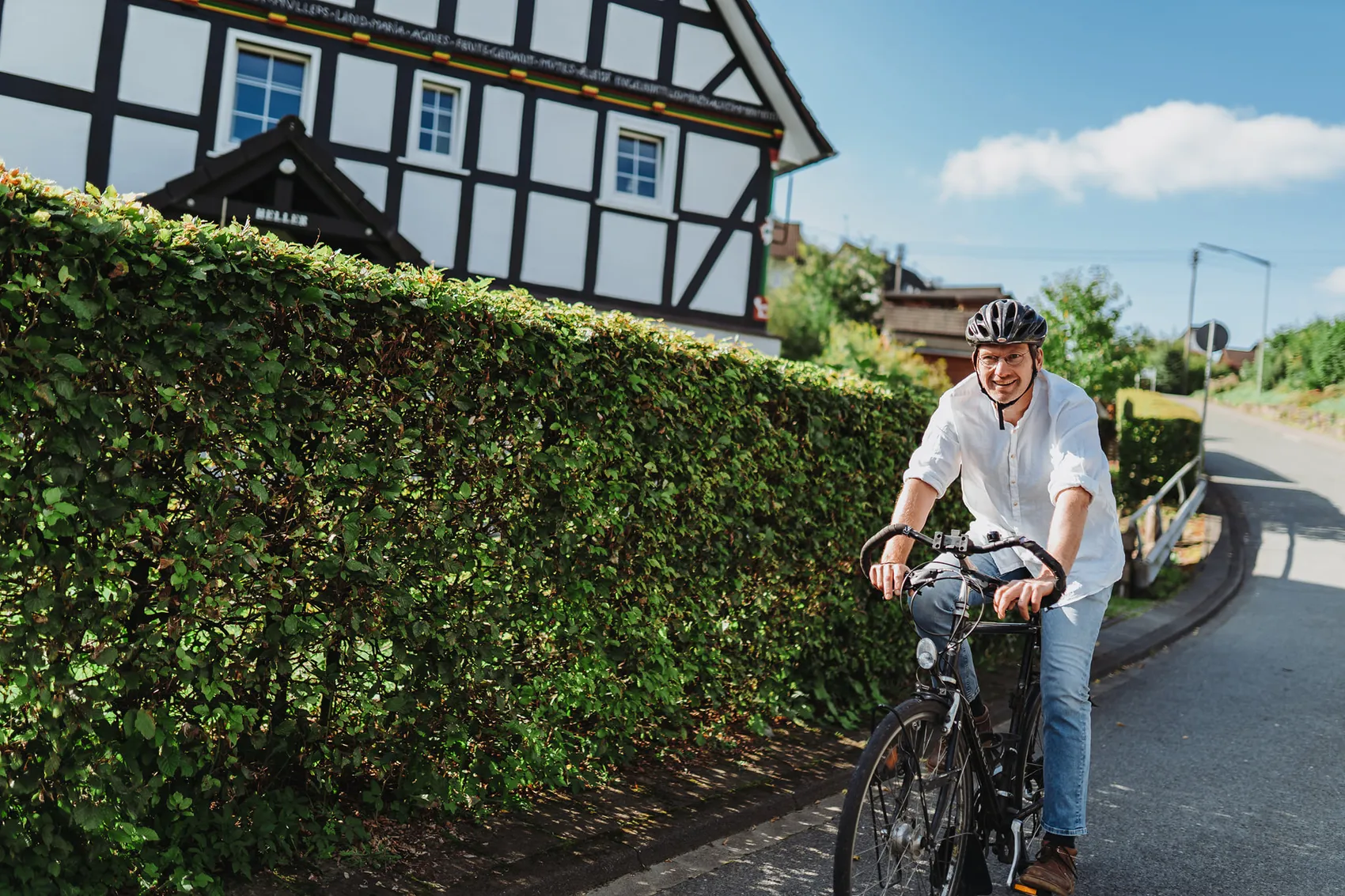 Gregor Kaiser, ein Mann mittleren Alters mit Helm, fährt auf einem Fahrrad auf einer Landstraße vor einem Fachwerkhause.