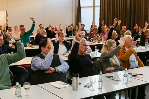 Bei einer Versammlung erheben viele Teilnehmende gleichzeitig die Hand zur Abstimmung. Tische mit Gläsern und Unterlagen stehen im Raum.