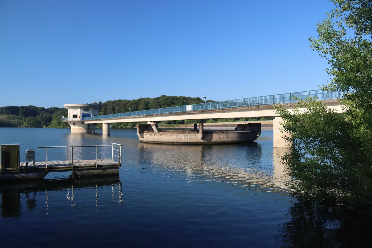 Eine Brücke mit Geländern überspannt das Wasser der Großen Dhünn-Talsperre. Links ein kleiner Steg, im Hintergrund Bäume unter blauem Himmel.