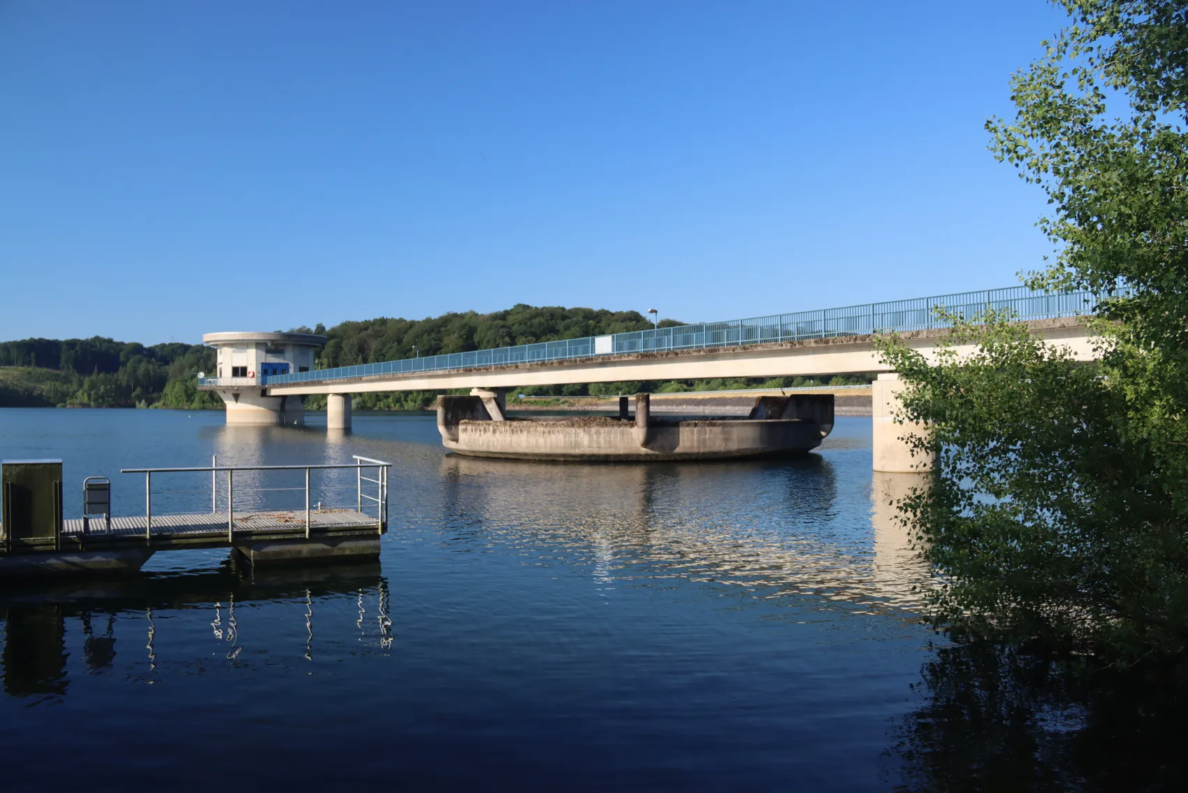 Eine Brücke mit Geländern überspannt das Wasser der Großen Dhünn-Talsperre. Links ein kleiner Steg, im Hintergrund Bäume unter blauem Himmel.