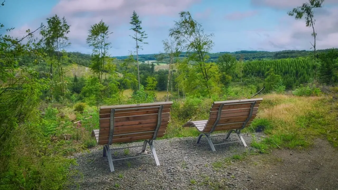 Zwei Holzliegen stehen auf einem Kiesweg mit Blick auf eine grüne Hügellandschaft und einen Wald.