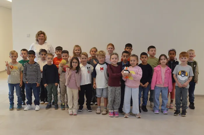 Eine Lehrerin und eine Gruppe von Grundschülern posieren für ein Klassenfoto vor einer weißen Wand.