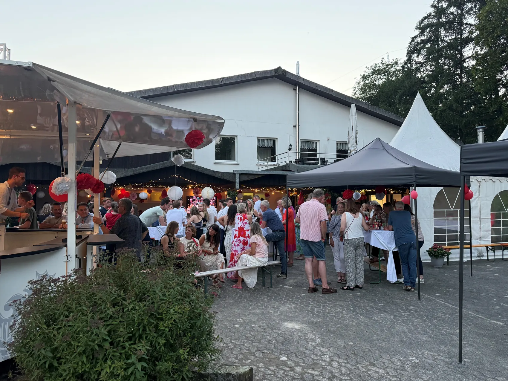 Menschen stehen und unterhalten sich auf einem Festgelände vor einem Gebäude und einem Foodtruck. Lampions und Girlanden schmücken den Veranstaltungsort.