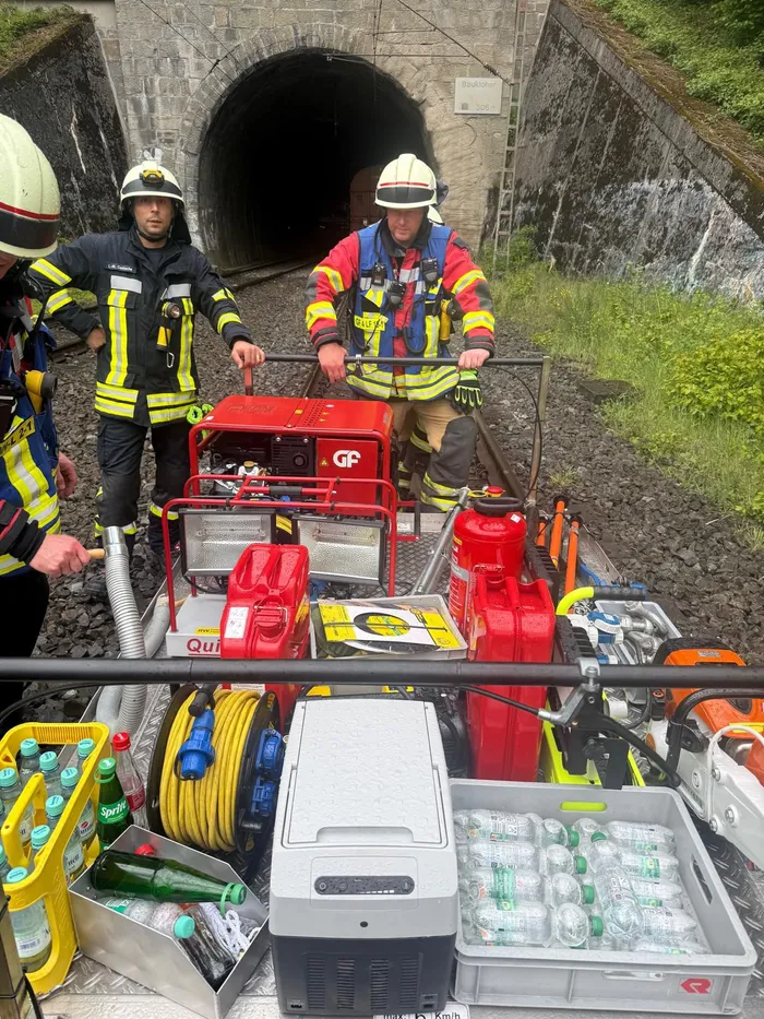 Mehrere Feuerwehrleute in Einsatzkleidung stehen vor einem roten Gerätewagen in einem dunklen Eisenbahntunnel. Vor dem Wagen sind Schläuche, Kisten mit Wasserflaschen und weitere Ausrüstung zu sehen.