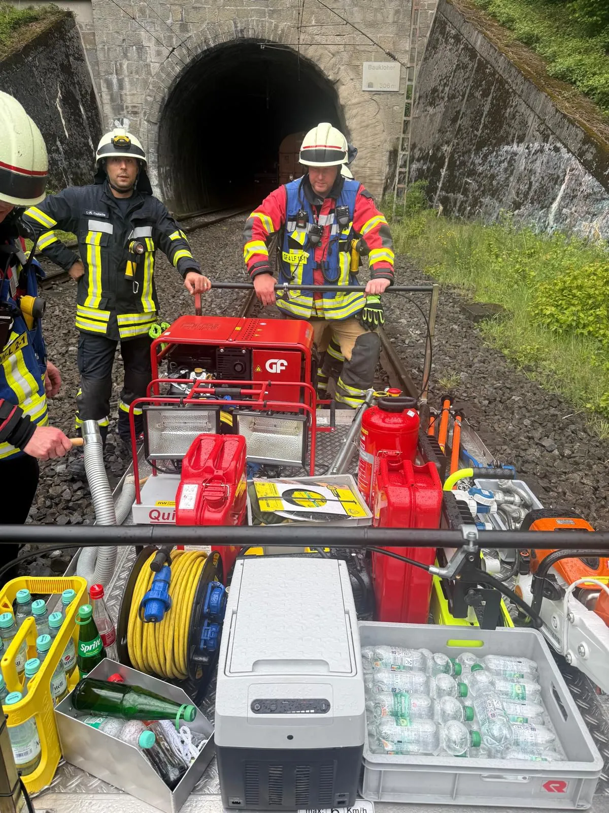 Mehrere Feuerwehrleute in Einsatzkleidung stehen vor einem roten Gerätewagen in einem dunklen Eisenbahntunnel. Vor dem Wagen sind Schläuche, Kisten mit Wasserflaschen und weitere Ausrüstung zu sehen.