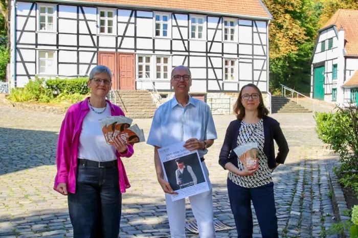 Gudrun Völcker, Dr. Thomas Eicher und Dr. Sandra Hertel halten Flyer und ein Plakat vor einem weißen Fachwerkhaus.