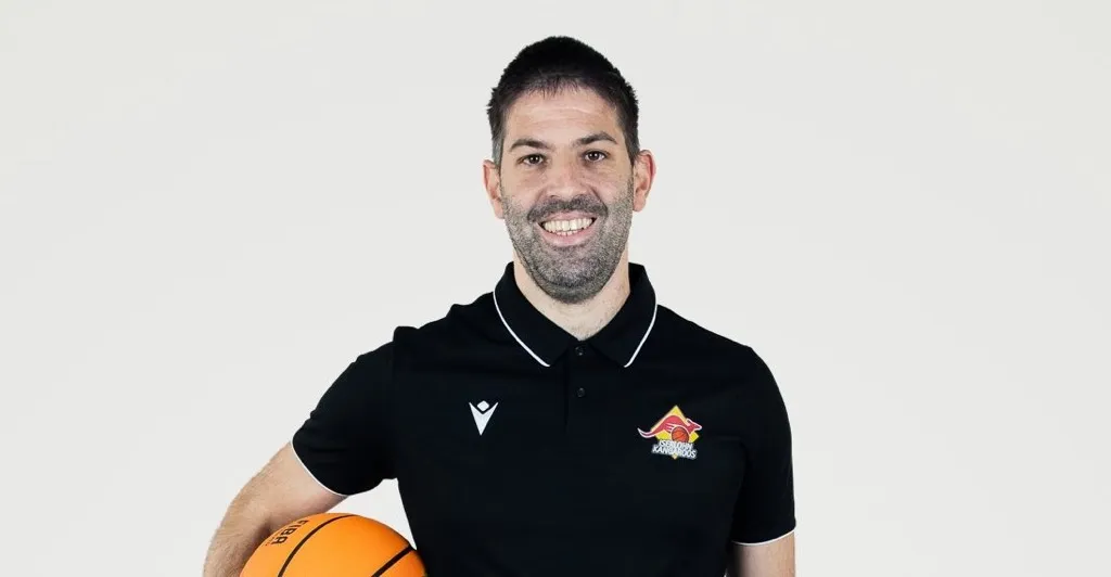 Lachender Mann mit kurzem dunklem Haar und Bart, trägt ein schwarzes Poloshirt mit dem Logo der Iserlohn Kangaroos und hält einen Basketball.