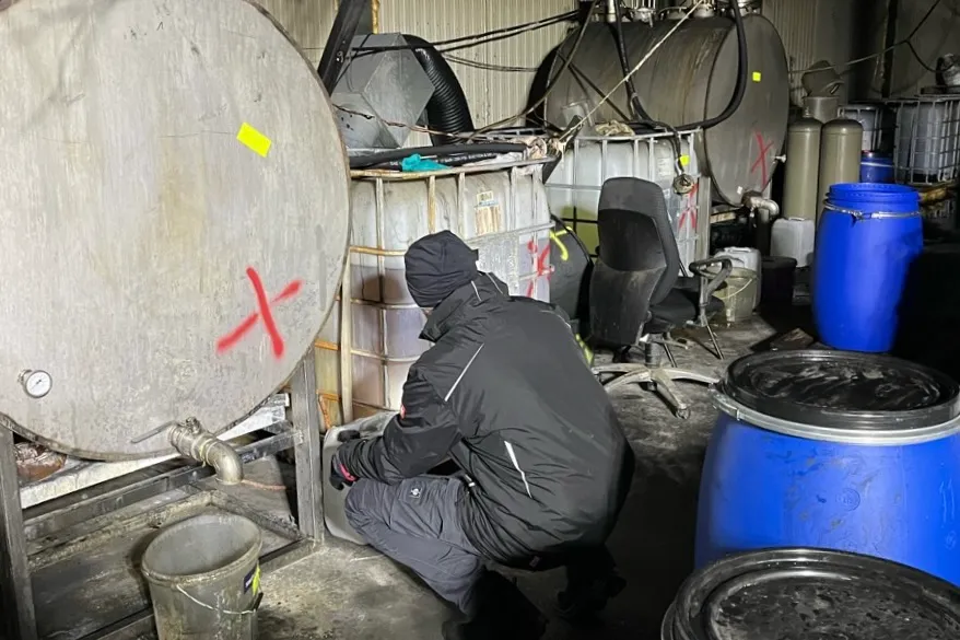 Eine Person in dunkler Kleidung hockt vor einem großen, metallenen Tank in einem Raum voller Behälter und Rohre.