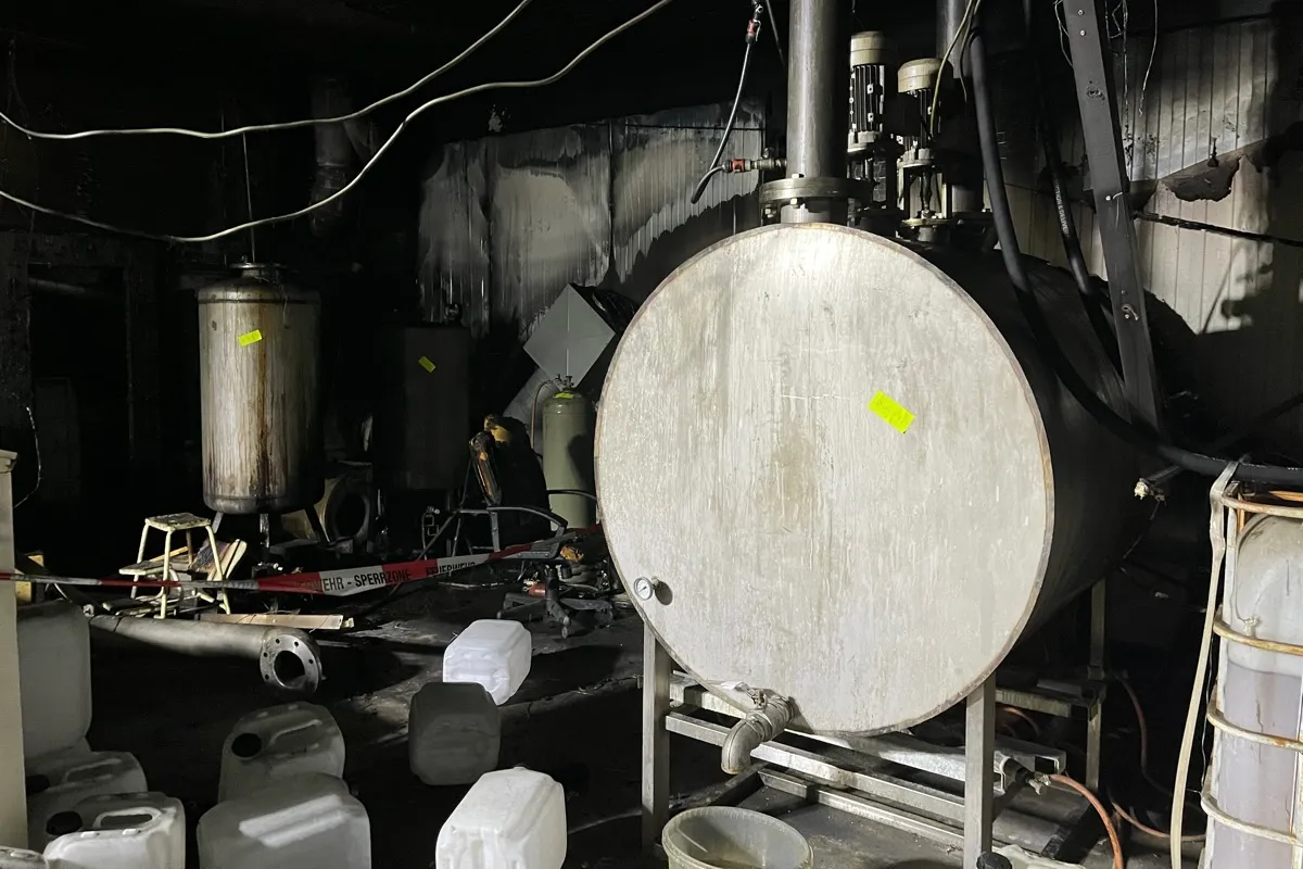 Mehrere metallene Reaktoren und Kanister stehen in einem dunklen Raum, der durch einen Brand geschwärzt wurde. Ein Warnschild mit der Aufschrift „GEFAHR – SPERRZONE“ ist zu sehen.