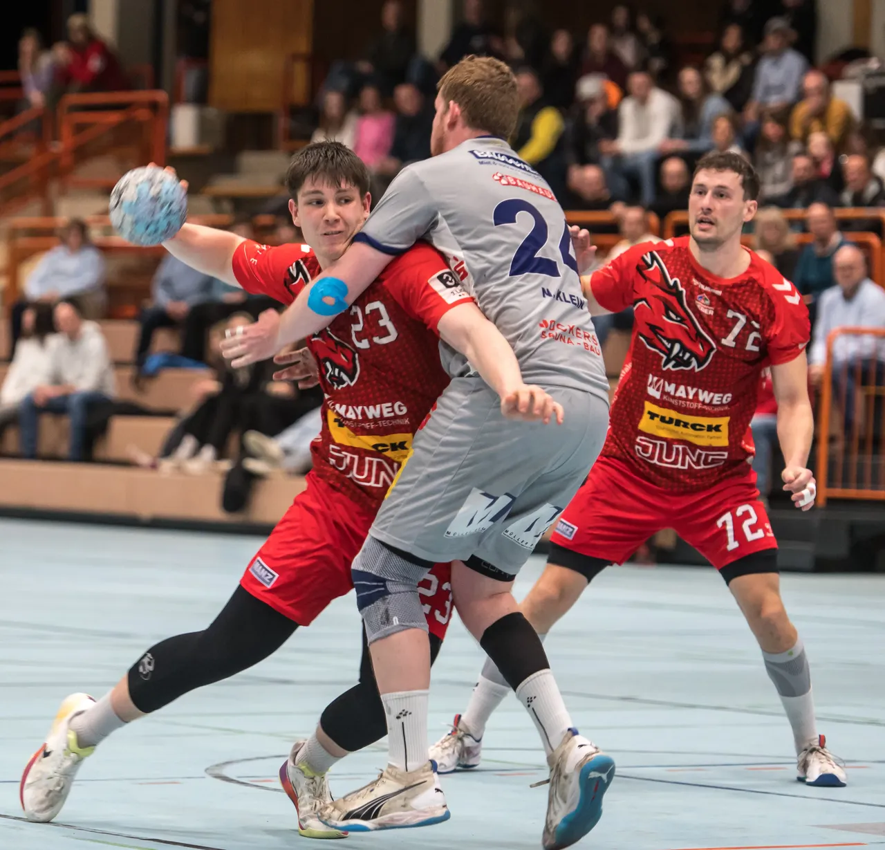 Zwei Handballspieler in roten Trikots versuchen, einen Spieler in grauen Shorts und Trikot mit der Nummer 2 zu blockieren. Im Hintergrund sind weitere Spieler und Zuschauer zu sehen.