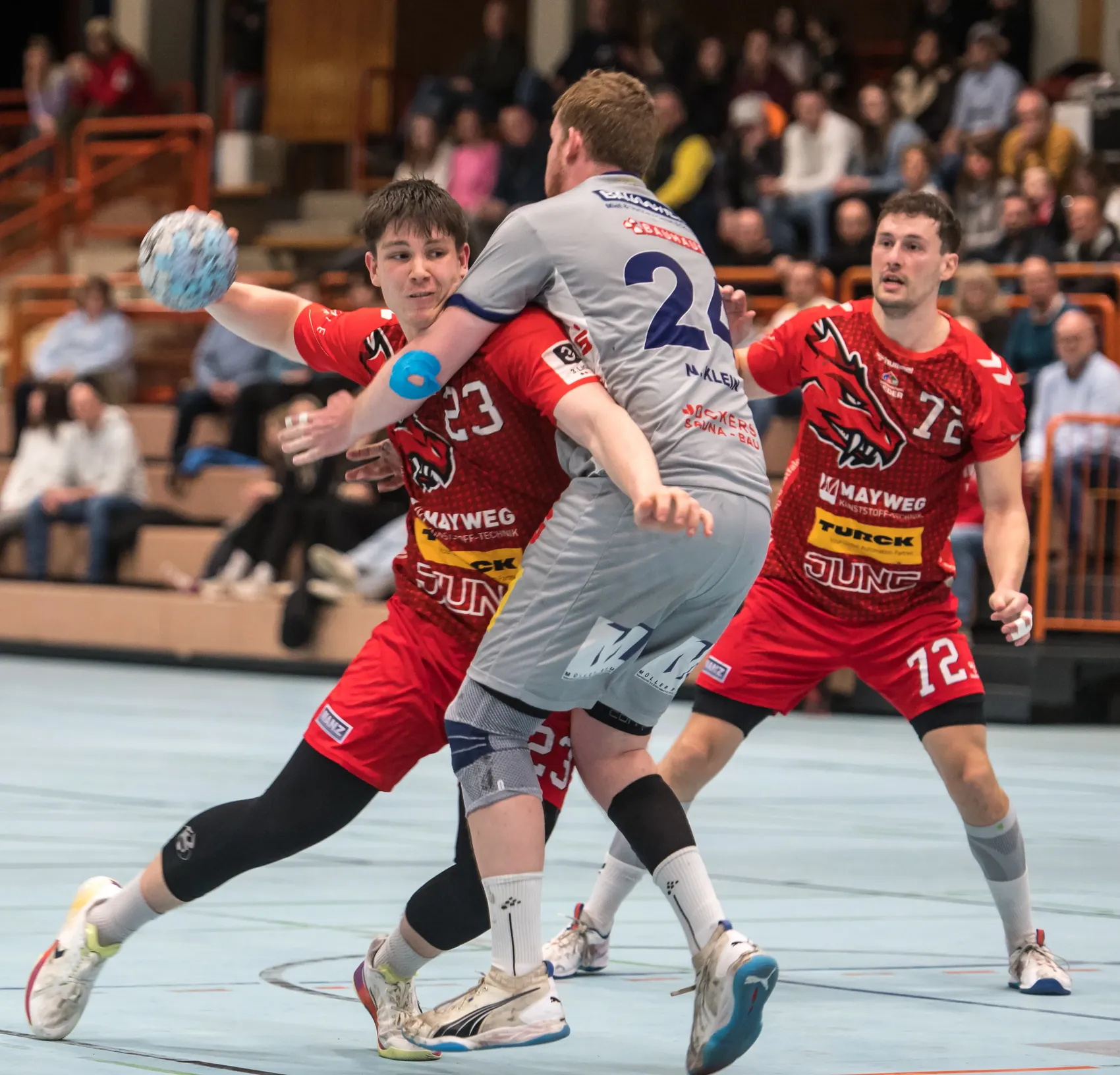 Zwei Handballspieler in roten Trikots versuchen, einen Spieler in grauen Shorts und Trikot mit der Nummer 2 zu blockieren. Im Hintergrund sind weitere Spieler und Zuschauer zu sehen.