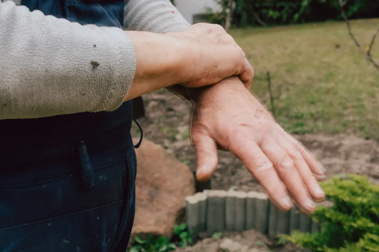 Eine Person hält sich mit einer Hand den Arm, die Hand zittert sichtbar. Im Hintergrund ein Garten.