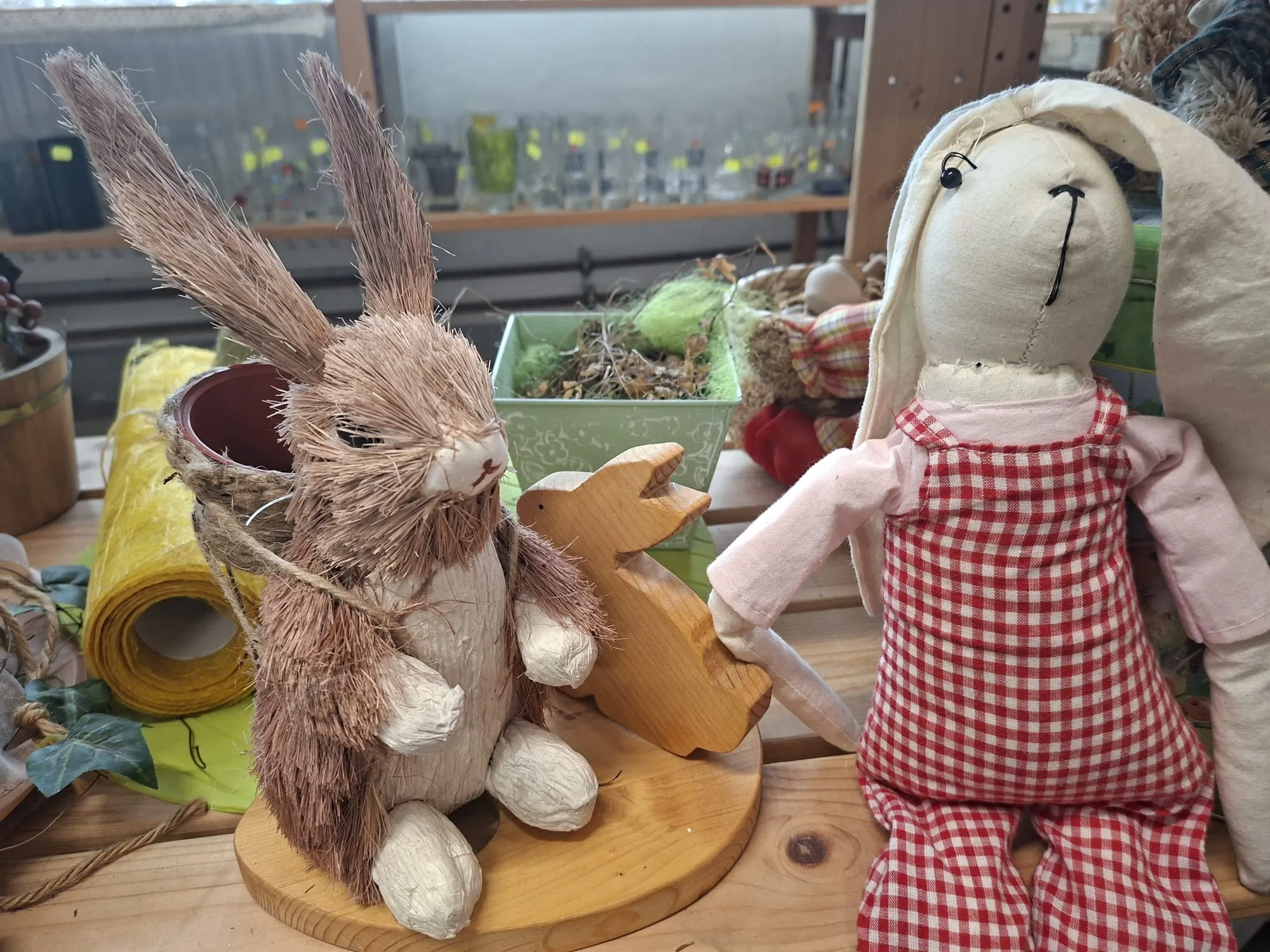 Zwei bekleidete Hasenfiguren aus Stoff und Stroh stehen vor einer verschwommenen Kulisse aus Pflanzen und Dekorationen.