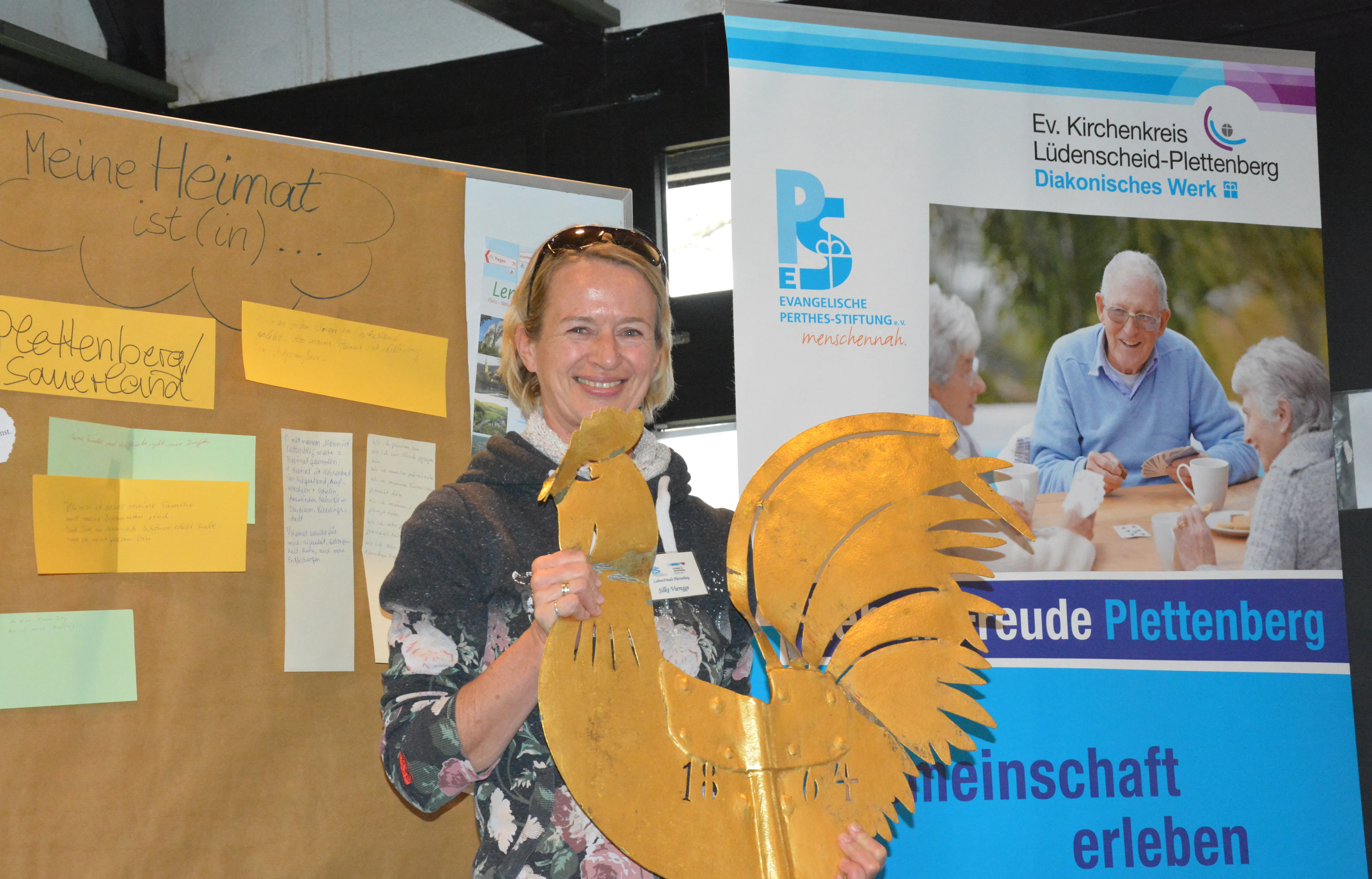 Moderatorin Silke Vieregge hält einen goldenen Hahn in der Hand, ein Symbol für Plettenberger Geschichte, vor einer Pinnwand mit handschriftlichen Beiträgen zum Thema Heimat.