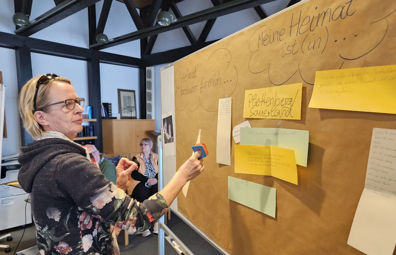 Eine Frau hängt mit einer Pinzette einen gelben Zettel an eine große Korkwand mit weiteren Notizen und Texten zum Thema Heimat.